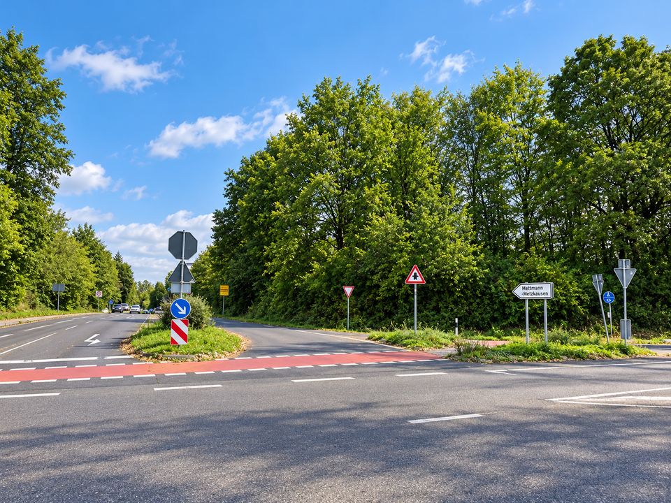 Die Kreuzung Ratinger Landstraße / Kantstraße an einem Sommertag: eine Verkehrsinsel mit Verkehrszeichen, ein rot markierter Radweg und ein Wegweiser nach Mettmann-Metzkausen, umgeben von hohen Bäumen.
